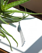 Silver pendant with tassels on a white surface next to a green plant