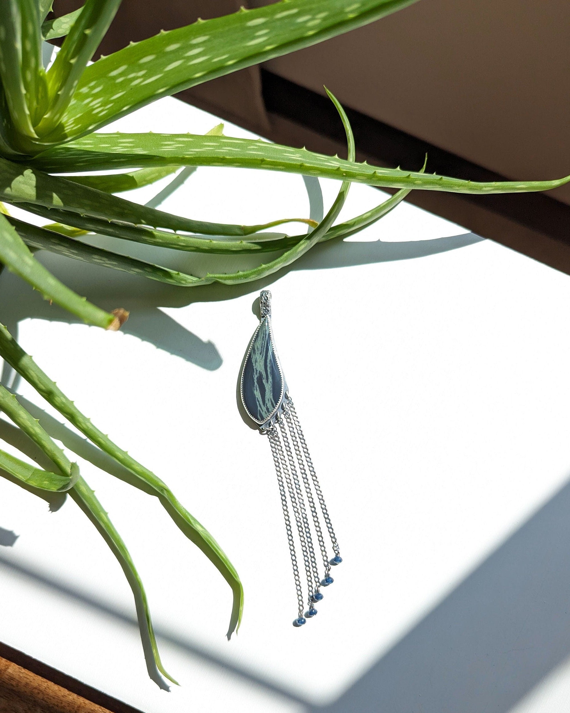 Silver pendant with tassels on a white surface next to a green plant