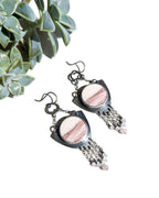 Pair of earrings with pink and mauve stones and grey metalwork against a white background with a plant.
