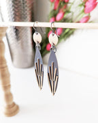 Pair of geometric earrings hanging on a rack with flowers in the background.