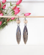 Pair of earrings with geometric design hanging on a stick with pink flowers in the background.