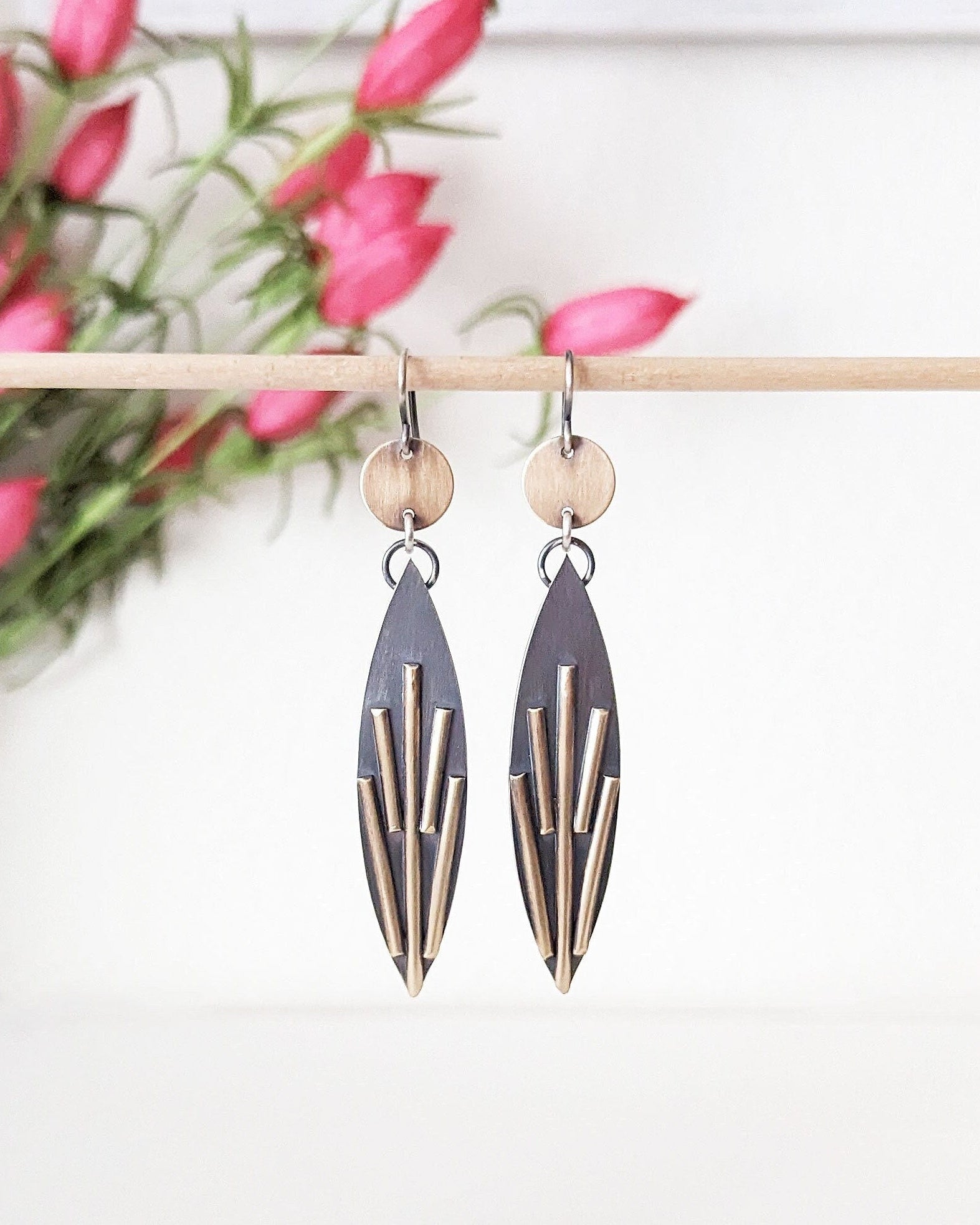 Pair of earrings with geometric design hanging on a stick with pink flowers in the background.
