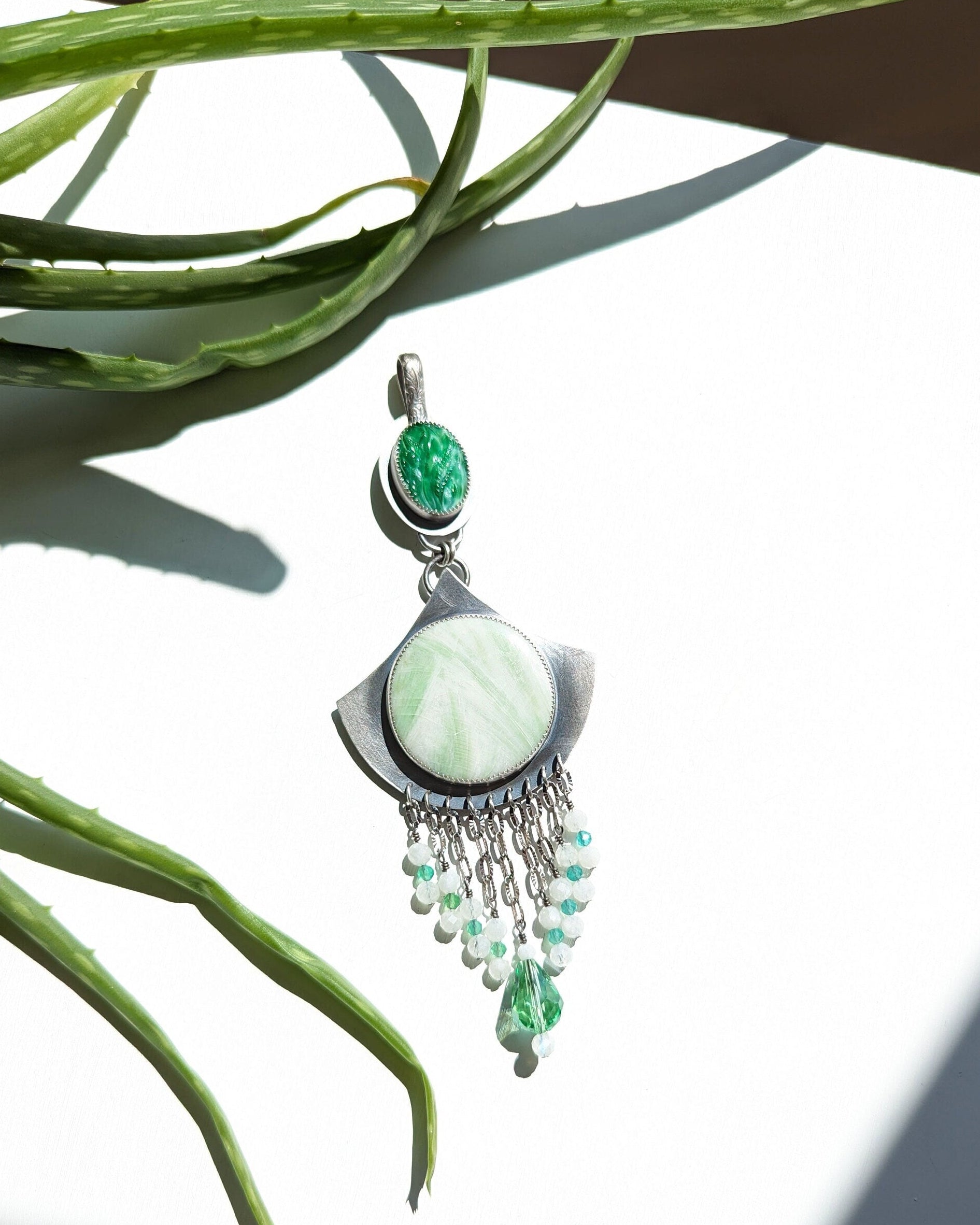 Green gemstone pendant with chain on a white surface with green leaves in the background