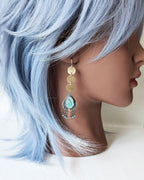 Mannequin head with blue hair and earrings on a white background, featuring long brass and turquoise fringe earrings.