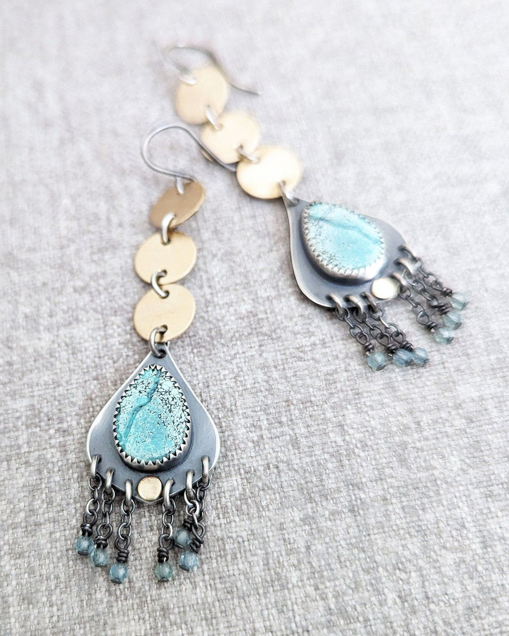 Pair of earrings with blue teardrop and gold circular elements on a gray fabric background.