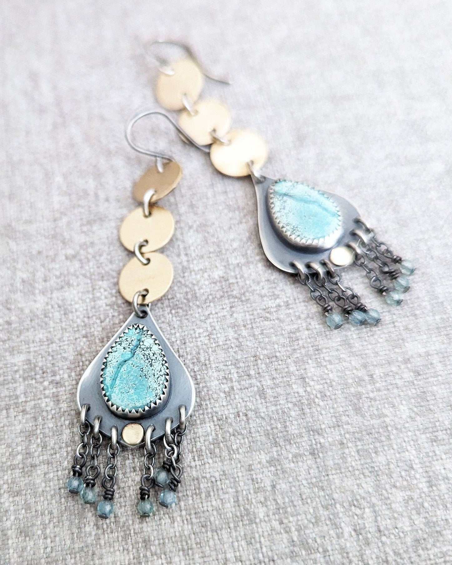 Pair of earrings with blue teardrop and gold circular elements on a gray fabric background.