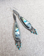 Pair of earrings with feather-like designs and turquoise beads on a textured surface