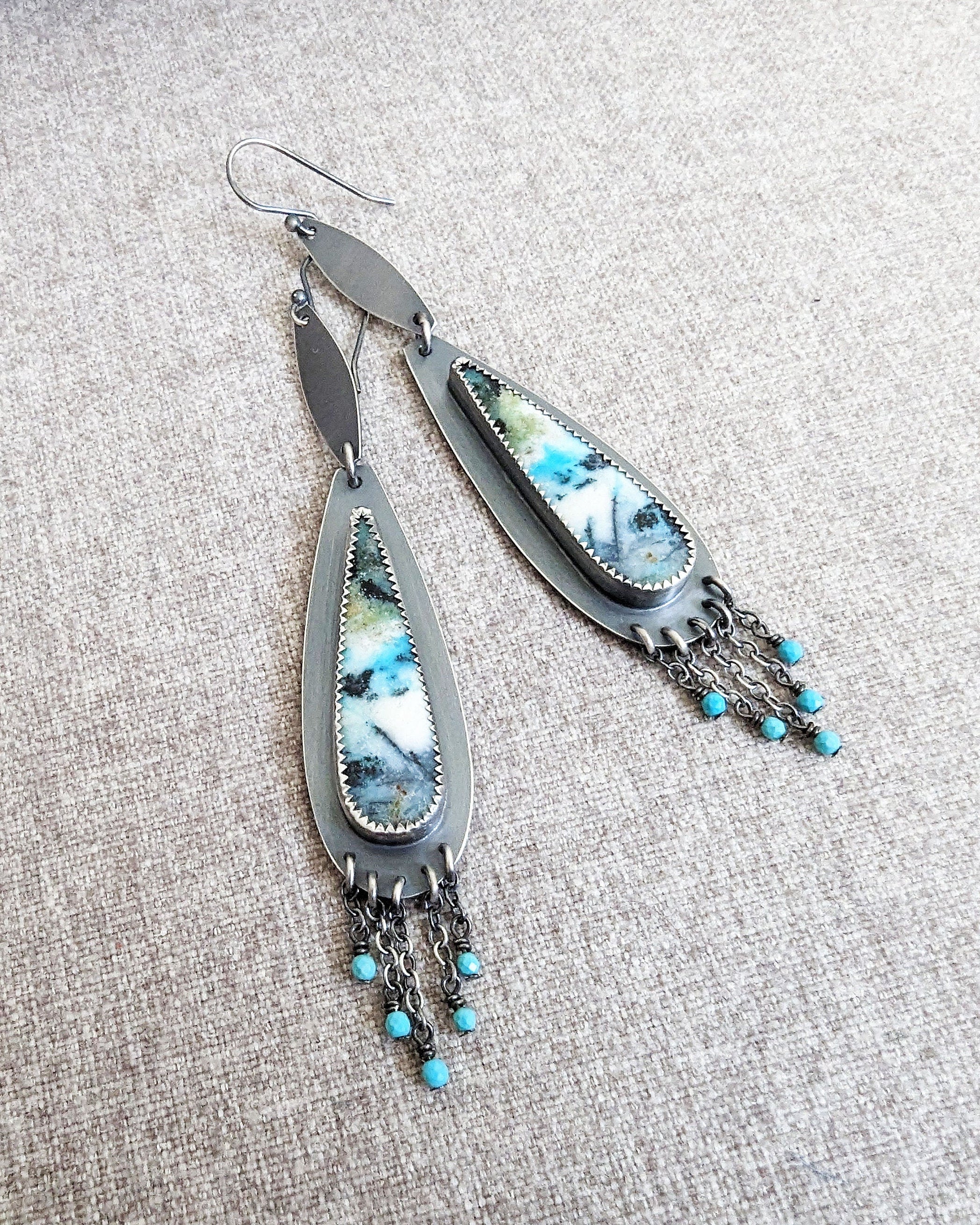 Pair of earrings with feather-like designs and turquoise beads on a textured surface
